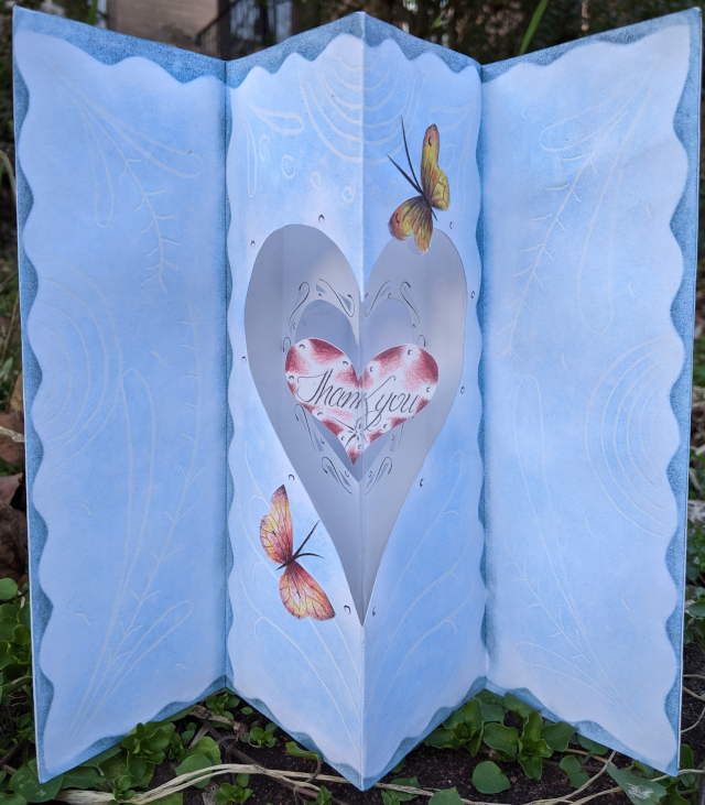[Pleated card showing butterflies approaching a heart-shaped plastic window in which a smaller heart is hanging by a thread. Written on the smaller heart are the words 'Thank you'.]